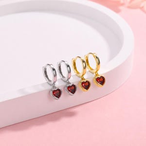 May include: Two pairs of hoop earrings, one pair in silver-tone and one pair in gold-tone metal. Each hoop features a small, dangling heart-shaped charm set with a deep red gemstone. The earrings are arranged in a row on a white circular base.