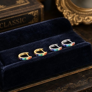 May include: Four small hoop earrings arranged in a row. Two earrings are gold-toned and two are silver-toned. Each hoop features a series of small, round, multi-colored gemstones set along the front edge, displaying a vibrant rainbow color gradient.