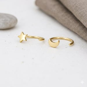 May include: Two gold-colored hoop earrings. One earring features a star design, and the other has a crescent moon. The earrings are set against a white surface with a neutral-colored fabric in the background.