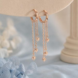 May include: A pair of rose gold hoop earrings featuring multiple dangling chains of varying lengths. Each chain is accented with a small, multi-pointed star charm. The earrings are constructed with a hinged closure mechanism for secure attachment.