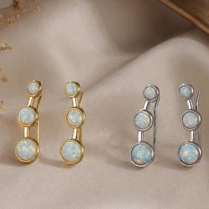 May include: Two pairs of ear climber earrings, one pair in gold-tone metal and one pair in silver-tone metal. Each earring features three round, iridescent white opal-style stones arranged in a vertical line, increasing in size from top to bottom.