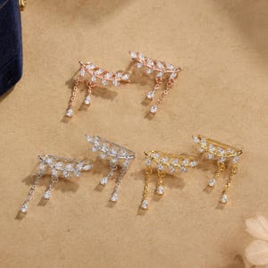 May include: Three pairs of ear climbers in rose gold, silver, and gold tones. Each earring features a curved branch design encrusted with clear marquise-cut crystals and two delicate dangling chains ending in small pear-shaped crystal drops.