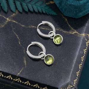 May include: A pair of silver hoop earrings, each featuring a small, round, translucent light green gemstone charm suspended from the bottom of the hoop. The metal has a polished finish, and the stones are smooth and circular in shape.