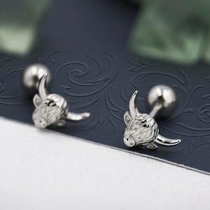 May include: A pair of silver-toned stud earrings featuring a detailed Highland cow head design. Each earring has a polished metallic finish with engraved textures representing the animal's shaggy coat and horns, secured with a round ball backing.