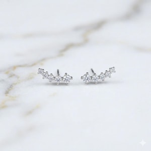 May include: A pair of silver-toned stud earrings. Each earring features a curved row of five clear, round-cut gemstones. The earrings are displayed on a white marble surface, showcasing their sparkle and design.
