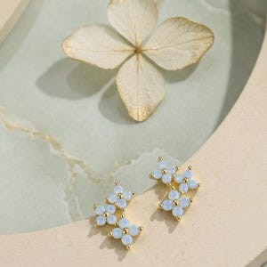 May include: Gold-toned stud earrings with a cluster of light blue, opal-like stones arranged in a floral design. The earrings are set against a soft, neutral background with a dried flower.