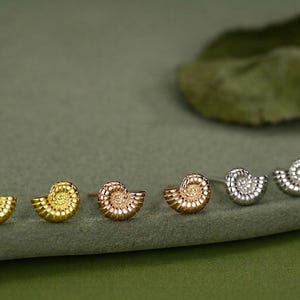 May include: A set of six small stud earrings shaped like spiraled ammonite shells. The collection includes two gold-toned, two rose gold-toned, and two silver-toned earrings, each featuring a detailed, textured surface with a classic spiral pattern.