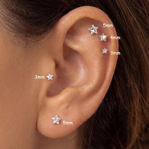 Pode incluir: Cinco brincos de pino em forma de estrela de tom prateado são usados em várias perfurações da orelha. Os brincos estão etiquetados com os seus tamanhos: 5mm, 4mm e 3mm. Cada estrela apresenta uma pedra central clara e facetada.