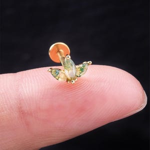 May include: A gold-colored stud earring with a floral design. The earring features three marquise-cut, light green gemstones with moss-like inclusions. The earring post is gold-colored, and the setting appears to be gold as well. The background is black.