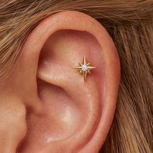 Pode incluir: Um brinco dourado em forma de estrela de quatro pontas com raios alongados e uma única pedra preciosa redonda e transparente cravada no centro. A joia está posicionada na área da cartilagem superior da orelha, exibindo um acabamento metálico.