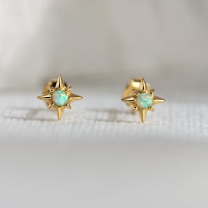 May include: A pair of gold-coloured stud earrings. Each earring has a starburst design with pointed edges and a central, round, opalescent stone. The earrings are set against a soft, white background.