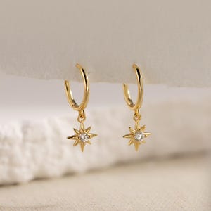 Könnte beinhalten: Ein Paar goldfarbene Creolen. Jeder Ohrring hat einen sternförmigen Anhänger mit einem kleinen, klaren Edelstein in der Mitte. Die Ohrringe werden vor einem weichen, neutralen Hintergrund präsentiert, der ihr zartes Design hervorhebt.