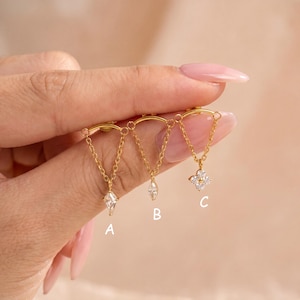 May include: Three gold-toned stud earrings featuring a curved bar connected to a delicate chain that suspends a small charm. Option A has a kite-shaped crystal, B has a marquise-cut crystal, and C has a four-petaled flower-shaped crystal.