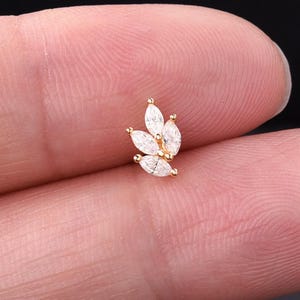 May include: A gold-colored earring with a cluster of five marquise-cut, clear gemstones. The gemstones are arranged in a leaf-like pattern, with small gold beads at the base of each gemstone. The earring is small and delicate.