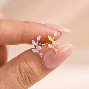 May include: Two stud earrings shaped like leafy branches, one in silver tone and one in gold tone. Each earring features clear marquise-cut stones arranged as leaves along a curved stem, with a single round brilliant-cut stone at the base.