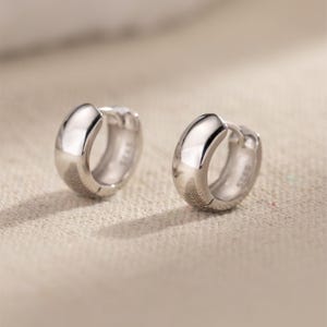 May include: A pair of small, shiny silver hoop earrings. The earrings have a rounded, polished design and a secure clasp. The jewelry is displayed on a neutral-colored surface, highlighting the reflective quality of the metal.