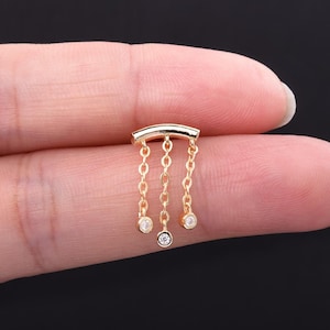 Puede incluir: Un pendiente dorado con un diseño de barra curva y tres cadenas colgantes. Cada cadena termina con una pequeña gema redonda y transparente. El pendiente se sostiene entre dos dedos sobre un fondo oscuro.