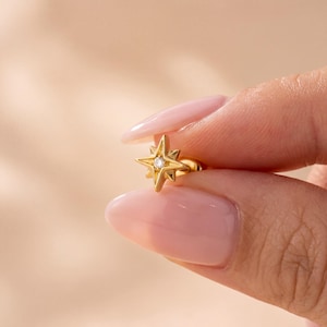 Pode incluir: Um brinco de pino dourado em forma de estrela de quatro pontas com uma pequena pedra preciosa redonda e transparente cravada no centro. A estrela apresenta pontas geométricas afiadas e um acabamento metálico polido que reflete a luz.