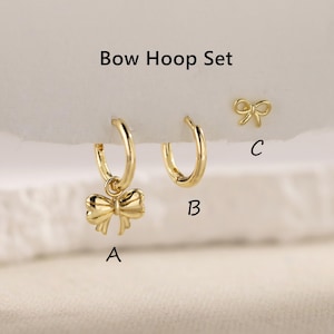 Puede incluir: Un conjunto de pendientes de aro dorados con forma de lazo, que incluye dos pendientes de aro y unos pendientes de botón con forma de lazo. Un pendiente de aro tiene un dije de lazo. El texto "Bow Hoop Set" está en la parte superior.