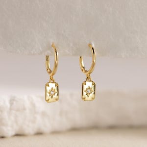 May include: A pair of gold-colored hoop earrings. Each earring features a rectangular charm with a starburst design and a small, clear gemstone in the center. The earrings are displayed against a soft, white background.