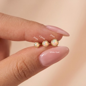 Pode incluir: Três brincos de opala branca redonda em engastes de ouro, rotulados com os tamanhos 2mm, 2,5mm e 3mm. As pedras exibem flashes iridescentes de cor e o texto White Opal está impresso abaixo para identificar o tipo de pedra preciosa.