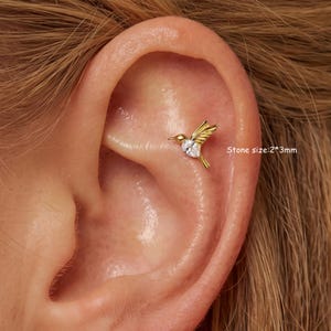 May include: A gold-toned hummingbird earring stud featuring a pear-shaped clear stone on the body. The earring is positioned in the upper ear cartilage. The text Stone size:2*3mm is visible next to the hummingbird design on the skin.