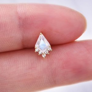 Pode incluir: Um pequeno brinco dourado com uma pedra preciosa iridescente em forma de losango. A pedra preciosa é rodeada por um grupo de pequenas pedras redondas e transparentes. O brinco é segurado entre dois dedos.