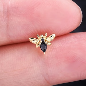 May include: A gold-colored bee-shaped earring with a black gemstone body. The wings and details of the bee are intricately designed. The earring is small and delicate, suitable for various ear piercings.