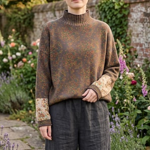 May include: A brown long-sleeve jumper with a ribbed mock neck collar and a repeating floral pattern in shades of orange, yellow, and green. The jumper features contrasting cuffs with a lighter floral print and is paired with dark grey trousers.