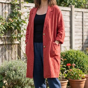 May include: A long, rust-coloured linen blazer with two large pockets and two brown buttons. The blazer is worn over a black top and blue trousers. The background includes a wooden fence, rose bushes, and potted plants.