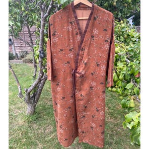 May include: A long, rust-coloured robe featuring a delicate floral pattern in muted tones. The garment has a V-neckline with dark brown trim, long sleeves, and a matching fabric tie closure at the waist. The fabric has a textured, woven appearance.