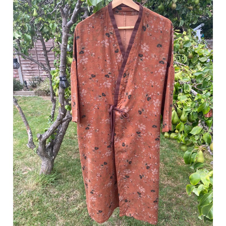 May include: A long, rust-coloured robe featuring a delicate floral pattern in muted tones. The garment has a V-neckline with dark brown trim, long sleeves, and a matching fabric tie closure at the waist. The fabric has a textured, woven appearance.
