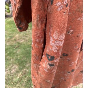 May include: A rust-coloured garment featuring a textured fabric with a repeating floral pattern in shades of muted pink, dark brown, and black. The item includes a visible side seam pocket and a short sleeve, showcasing the weave of the material.