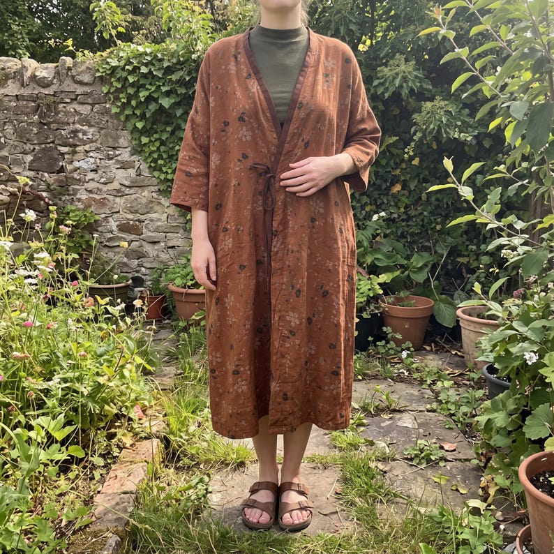 May include: A long, rust-coloured open-front robe featuring a subtle floral pattern in darker brown and grey tones. The garment has three-quarter length sleeves, a dark brown trim along the neckline, and a thin tie closure at the waist.