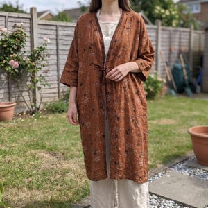May include: A long, open-front rust-coloured duster coat featuring a dark brown floral pattern, three-quarter length sleeves, and a thin tie closure at the waist. The garment is worn over a light-coloured, neutral-toned shirt and matching trousers.
