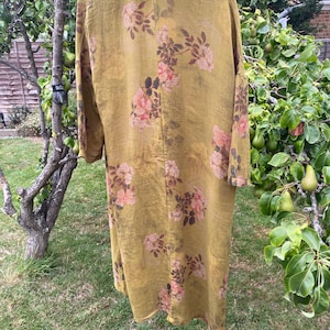 May include: A long-sleeved, mustard-yellow duster coat featuring a repeating floral pattern of pink roses and dark brown leaves. The garment has a dark brown collar and is displayed on a wooden hanger, showcasing a loose, flowing silhouette.
