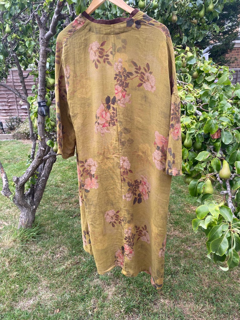 May include: A long-sleeved, mustard-yellow duster coat featuring a repeating floral pattern of pink roses and dark brown leaves. The garment has a dark brown collar and is displayed on a wooden hanger, showcasing a loose, flowing silhouette.