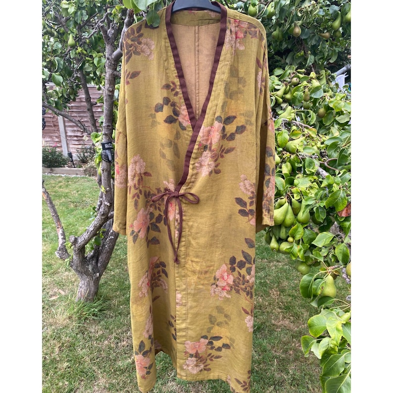 May include: A long, mustard-yellow robe featuring a floral pattern with muted pink and brown blossoms and leaves. The garment has a dark brown trim along the front opening and a matching fabric tie closure at the waist with long, loose sleeves.