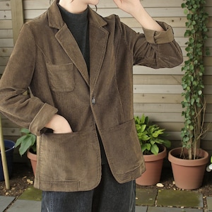 May include: A brown corduroy blazer with a notched lapel, a single-button closure, and two front pockets. The sleeves are cuffed, and the blazer is worn over a dark grey turtleneck. The blazer has a classic, tailored design.