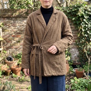 May include: A brown wrap-style jacket featuring long sleeves, a notched lapel collar, and a fabric tie closure at the waist. The garment is constructed with visible stitching along the edges and includes two front pockets, worn over a black shirt.