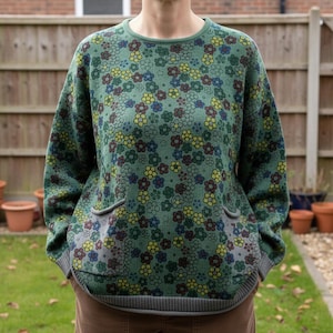 May include: A green floral patterned sweater with yellow, blue, and maroon flowers. The sweater features two front pockets and grey trim. The cuffs, hem, and pockets are trimmed in grey. The sweater is made of a knitted material.