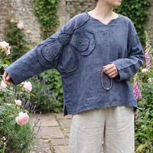 May include: A slate blue long-sleeve top featuring a large, dimensional fabric flower appliqué on the right shoulder and sleeve. The garment has a V-neckline, a relaxed fit, and side slits at the hem, paired with loose-fitting beige trousers.