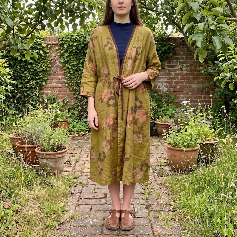 May include: An olive green duster coat featuring a floral pattern in shades of peach and brown. The garment has three-quarter length sleeves, a dark brown trim along the open front, and a thin brown tie closure at the waist over a dark blue shirt.