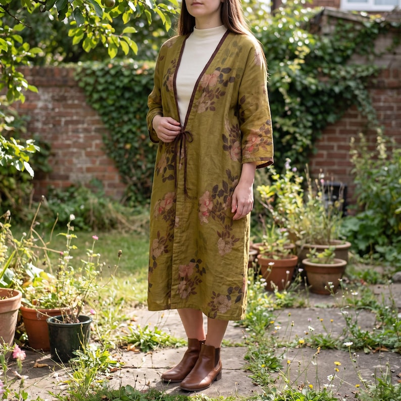 May include: A long, open-front olive green duster coat featuring a dark brown floral print, three-quarter length sleeves, and a thin tie closure at the waist, worn over a cream-coloured mock neck top and brown leather ankle boots.