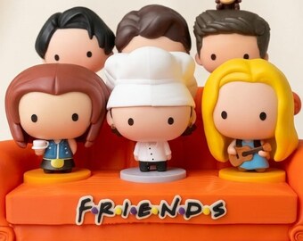 Può includere: Un set di figurine decorative con sei personaggi dei cartoni animati della serie TV "Friends" su un divano arancione. Il divano ha la scritta "FRIENDS" sul davanti. I personaggi hanno grandi teste e indossano vari abiti.