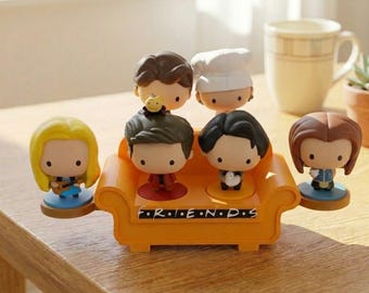 May include: A collection of six cartoon figurines of characters from the TV show Friends. The figurines are on a small orange couch with the show's logo. The characters have large heads and are on circular bases.