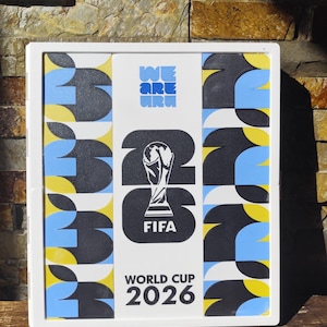 May include: A rectangular white frame containing a graphic display for the FIFA World Cup 2026. The center features the tournament trophy icon, the text WE ARE 26, FIFA, and WORLD CUP 2026. The side panels feature a repeating pattern of black, blue, and yellow.