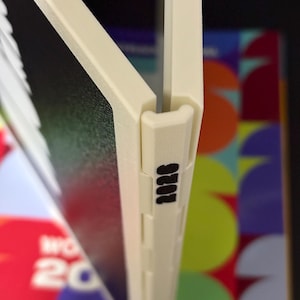 May include: A cream-colored plastic binder spine featuring a hinge mechanism and the black printed text 2026. The spine connects two dark panels, with a colorful graphic background visible behind the structure, showing partial text and shapes.