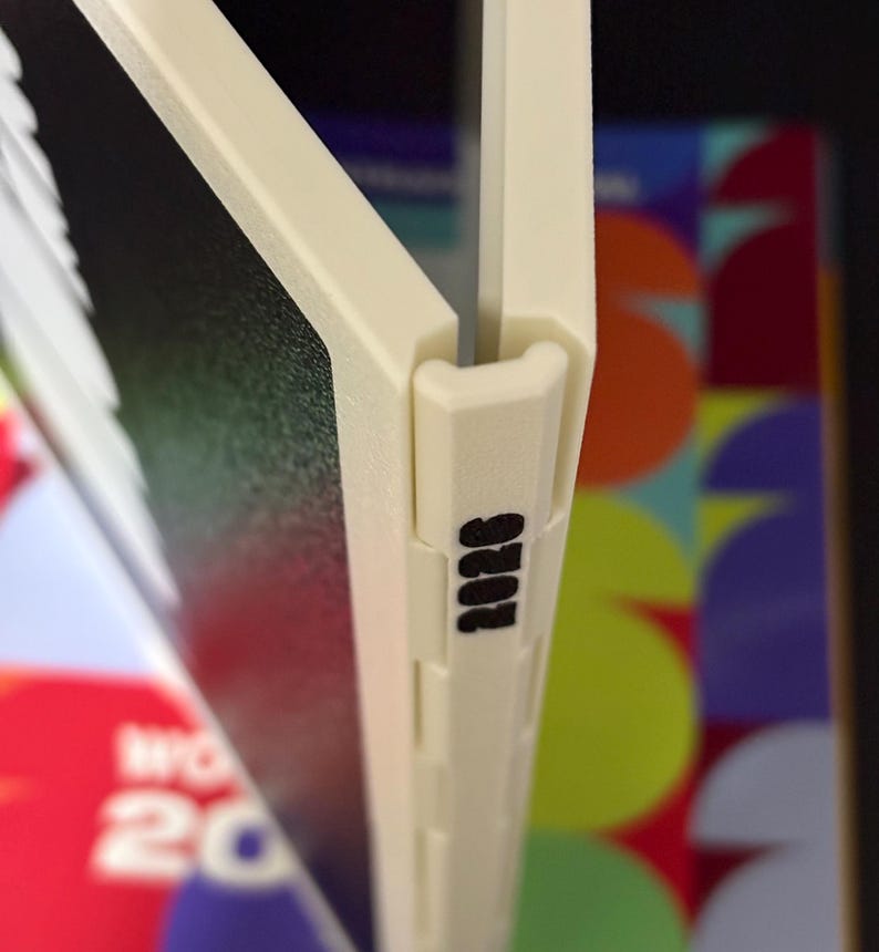 May include: A cream-colored plastic binder spine featuring a hinge mechanism and the black printed text 2026. The spine connects two dark panels, with a colorful graphic background visible behind the structure, showing partial text and shapes.