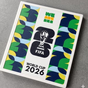 May include: A square white frame containing a graphic print for the FIFA World Cup 2026. The center features the trophy icon over the number 26, with the text WE ARE BRA, FIFA, and WORLD CUP 2026. The sides have a repeating pattern of green, blue, yellow, and light blue shapes.